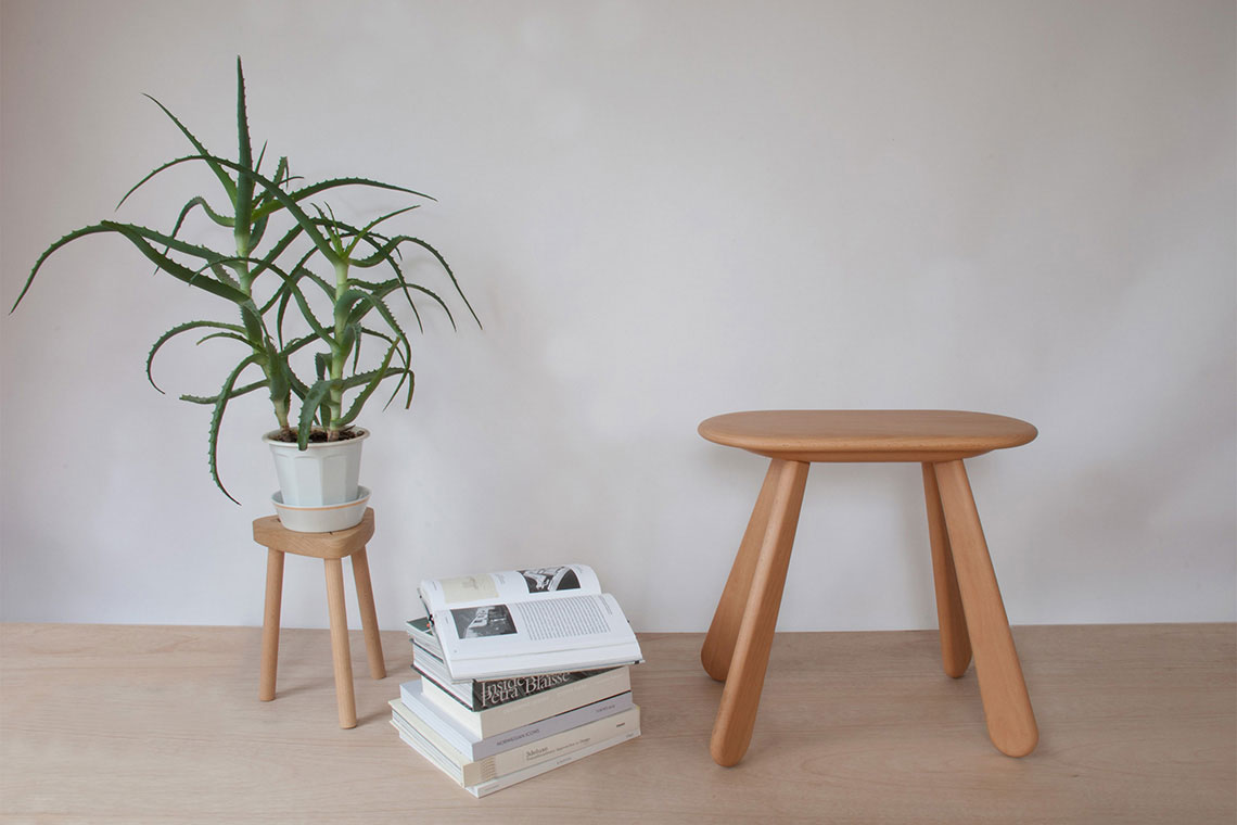 Solid beech wood stool with soft sculptural form designed by 24d-studio is shown in interior setting with green plant and stacked books.